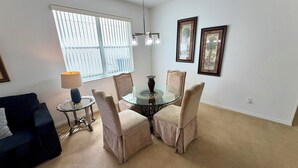 A dining area with a round glass table, four cushioned chairs, a small side table with a lamp, and a window with blinds.