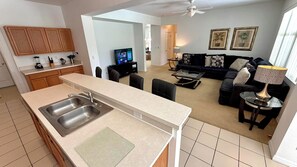 A modern kitchen with a large island, a dining area with a glass table, and a living room with a dark-colored sofa and decorative pillows.