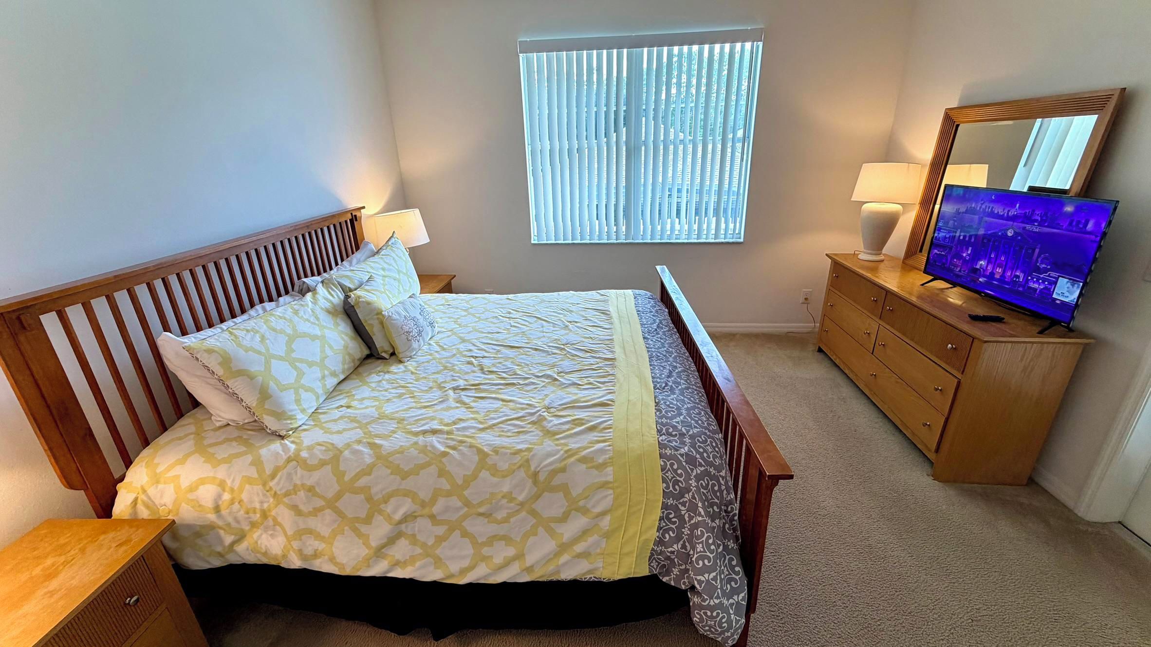A bedroom with a bed, a dresser, a television, and a window with blinds.