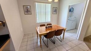 A dining area with a wooden table and chairs, a window with blinds, and a laundry room with a washing machine and a basket.