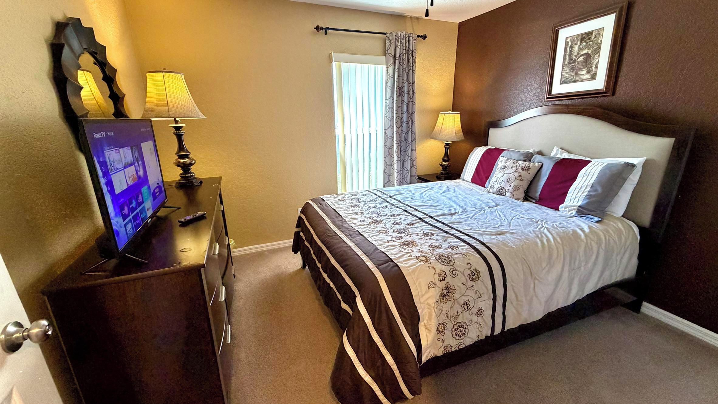 A hotel room with a bed, two bedside lamps, a television on a wooden stand, a framed picture on the wall, and a window with curtains.