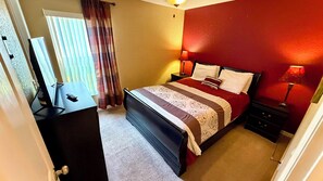 Un dormitorio con cama, lámparas de noche, una ventana con cortinas y un escritorio con control remoto.