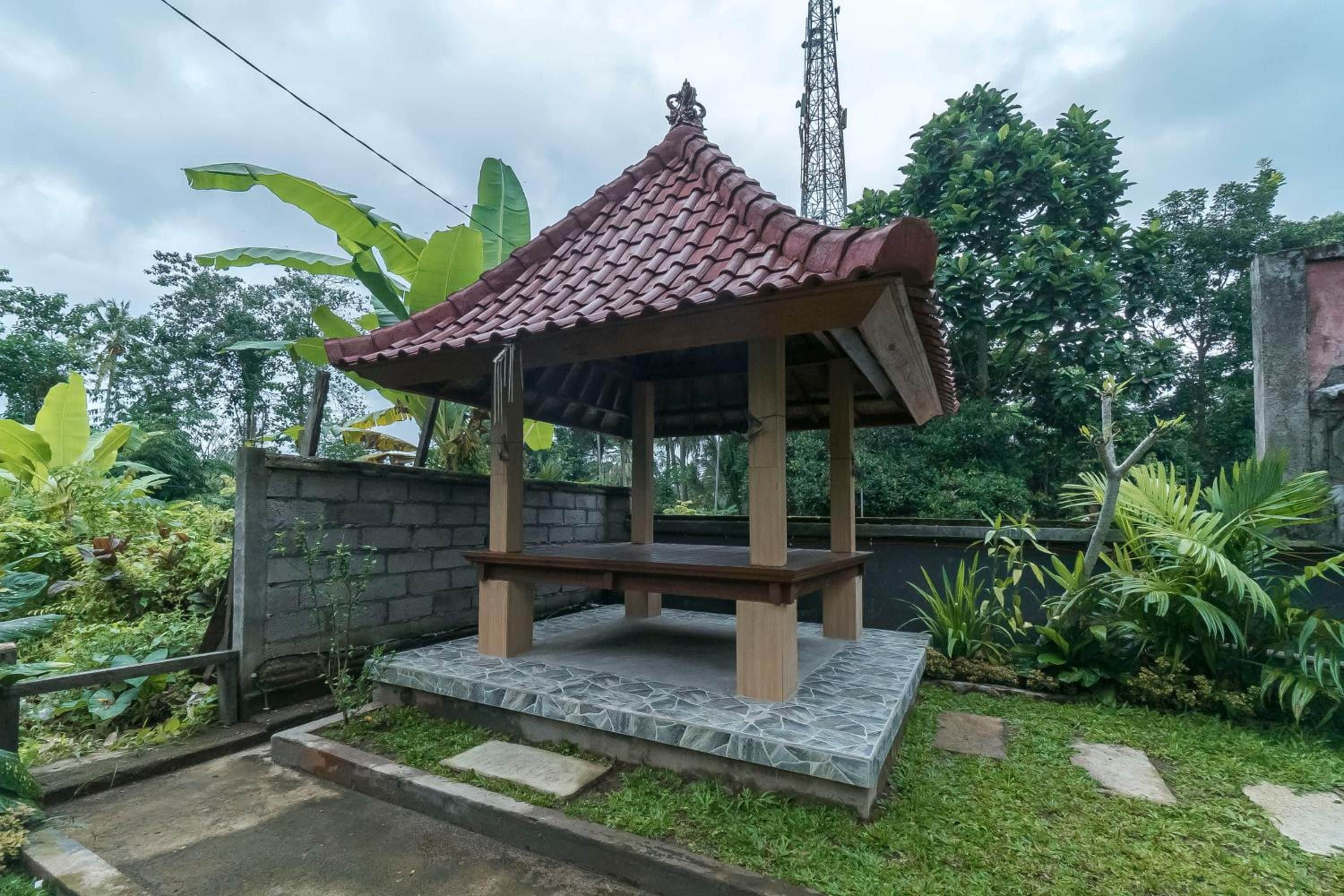 A gazebo with a tiled roof and a stone base, surrounded by lush greenery and a stone wall.