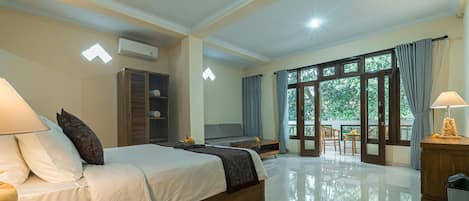 A hotel room with a bed, bedside table, lamp, wardrobe, and a view of greenery through large windows.