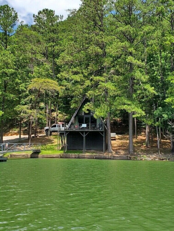 A cabin with a sloped roof, surrounded by dense trees, located near a body of water.