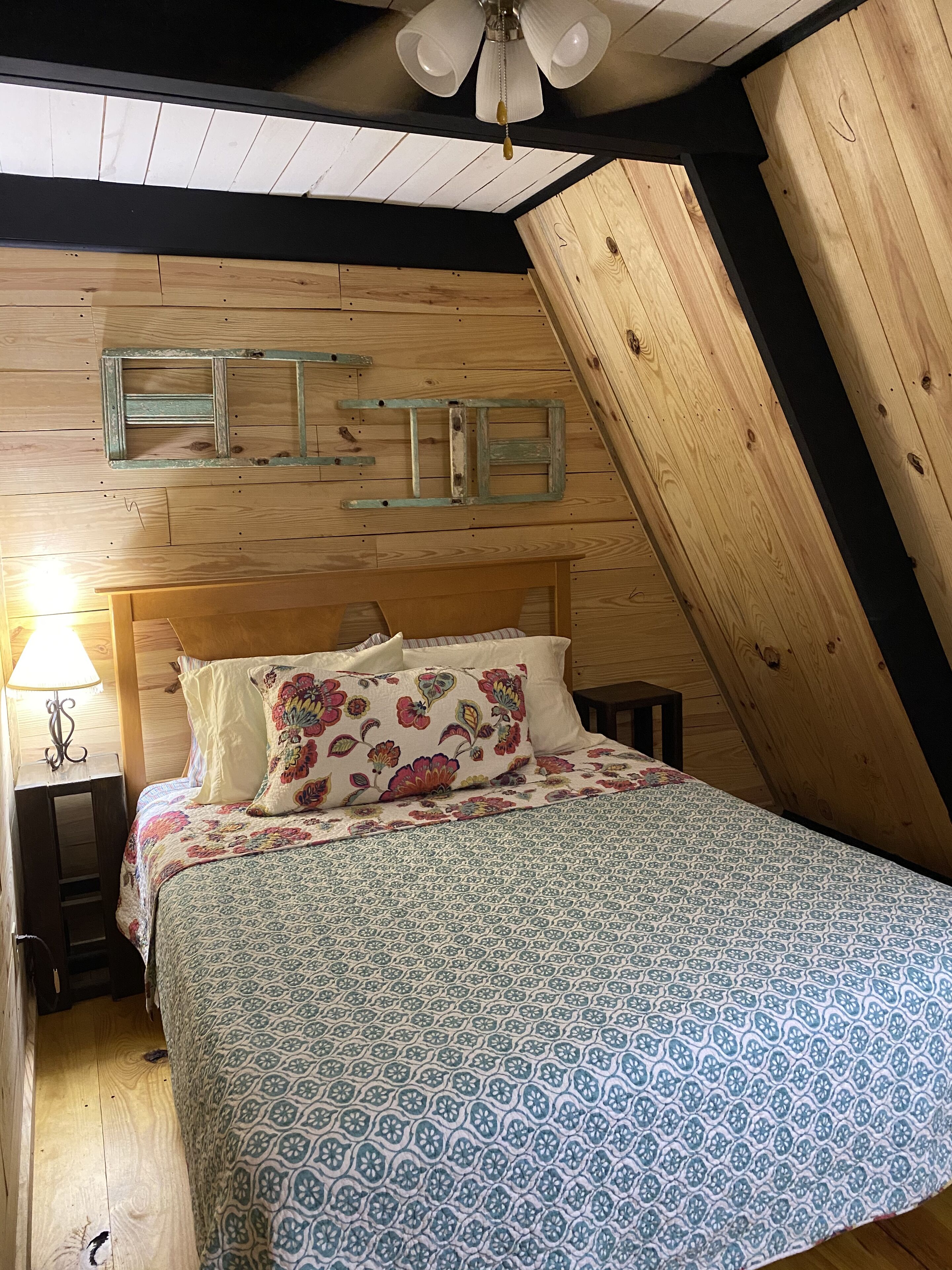 Ein gemütliches Schlafzimmer mit schrägen Wänden, Holzwänden, einem Bett mit Blumenmuster und einem Deckenventilator.
