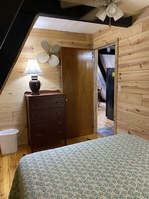 A bedroom with a sloped ceiling, a wooden dresser, a patterned bedspread, and a ceiling fan.