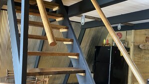 Eine Holztreppe mit Metallrahmen, die zu einem Küchenbereich mit einem schwarzen Kühlschrank und Holzsesseln führt.