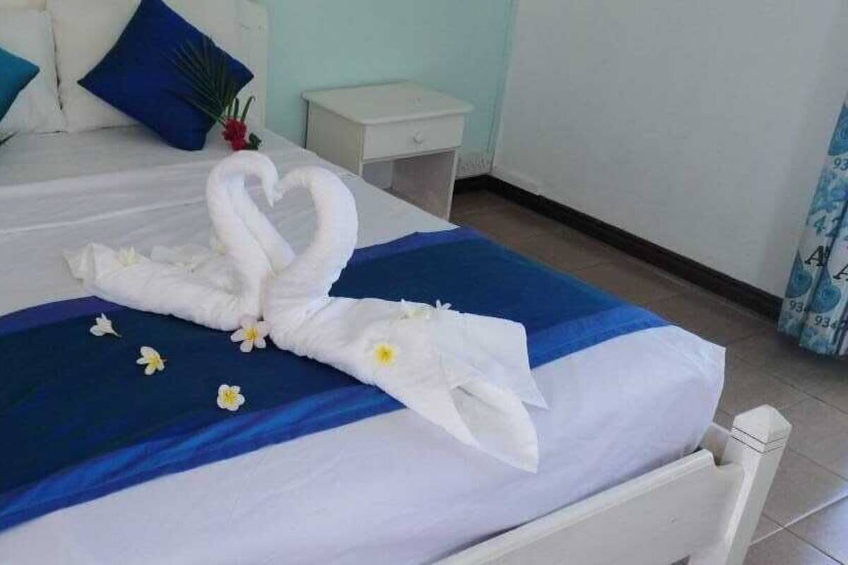 A neatly made bed with a blue bedspread and white linens, adorned with a swan-shaped towel and small flowers.