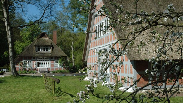 A traditional thatched-roof house with a brick facade, surrounded by a well-maintained garden and trees. - Atelier 2 - Atelierhaus Tannenweg (Worpswede)