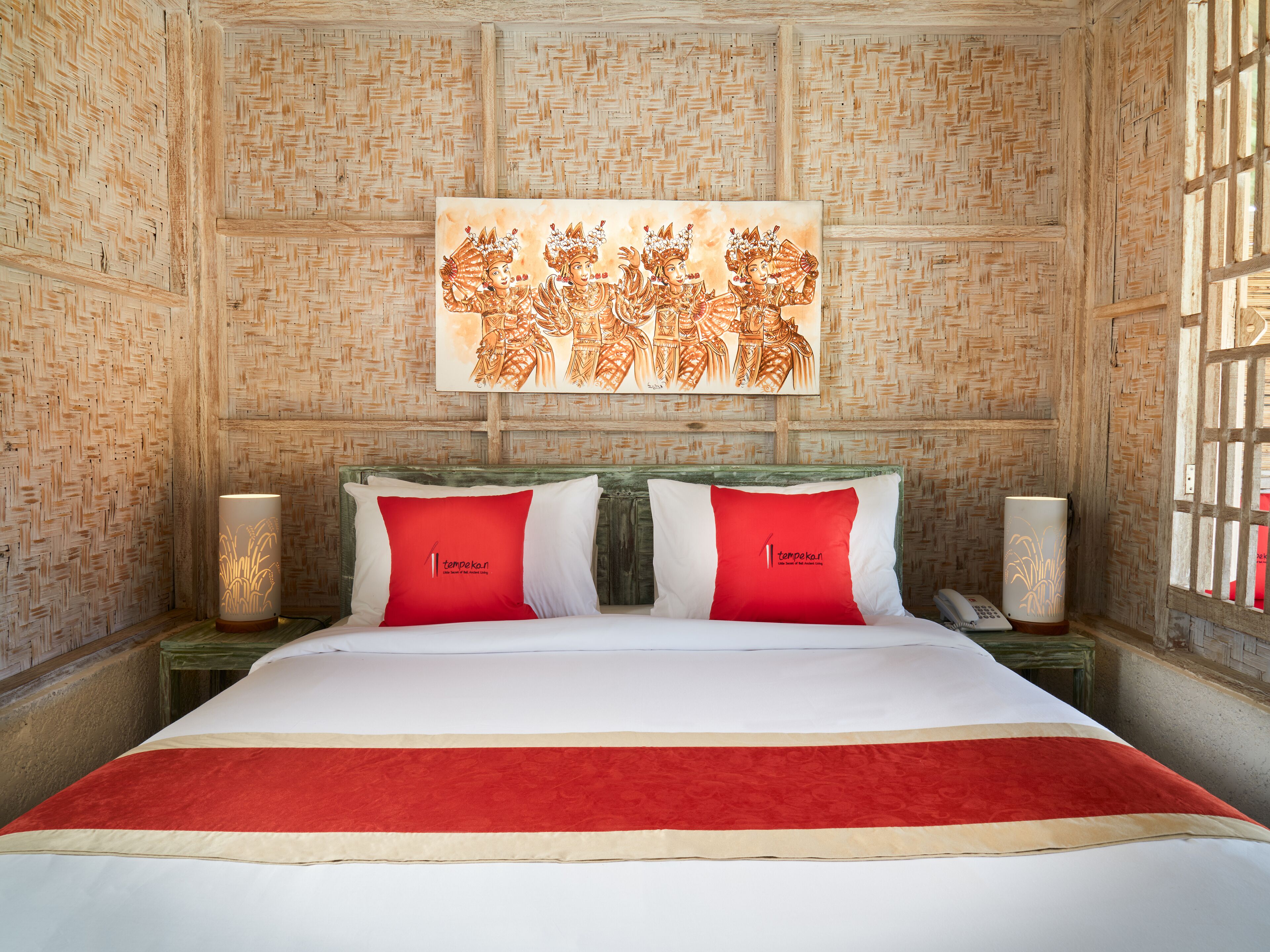 A hotel room with a bed, two red pillows, a wall tapestry, and two bedside lamps.
