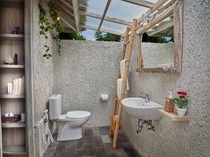 Un baño con inodoro, lavabo y zona de ducha, que incluye un estante de madera con artículos de aseo y un perchero para toallas.