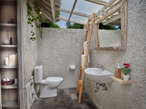 A bathroom with a toilet, sink, and shower area, featuring a wooden shelf with toiletries and a hanging towel rack.