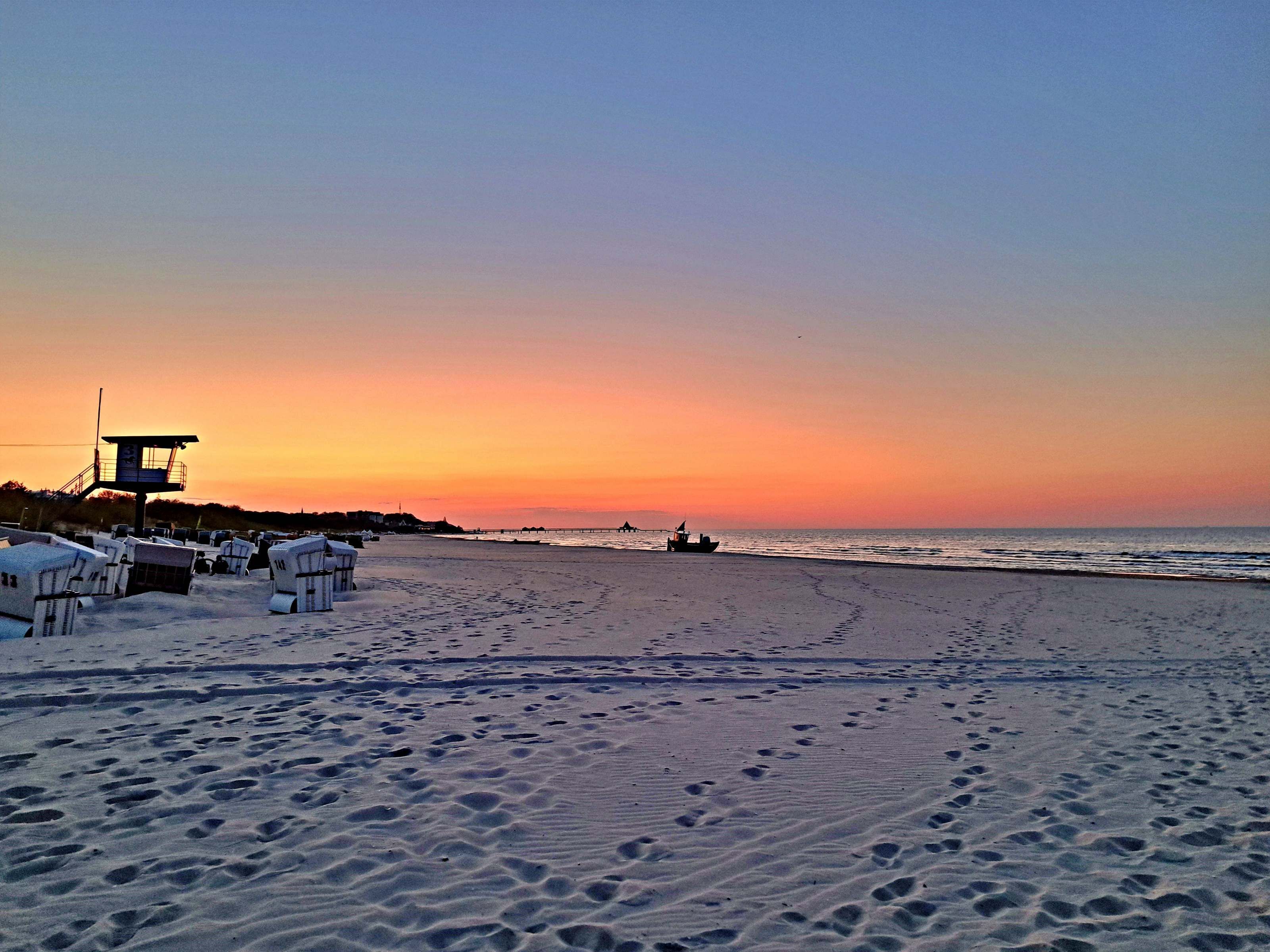 Ein Strand mit Fußspuren, einem Rettungsturm und Liegestühlen.