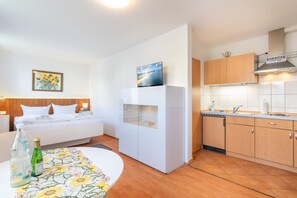 Ein kompaktes Hotelzimmer mit einem Bett, einem weißen Kleiderschrank, einer kleinen Küchenzeile mit Holzschränken und einem Esstisch mit blumigem Tischläufer.