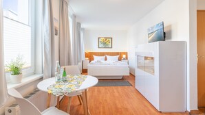 Habitación de hotel pequeña con cama, una mesita con mantel floral, refrigerador y televisor sobre un soporte.