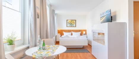 A compact hotel room with a bed, a small table with a floral tablecloth, a refrigerator, and a television on a stand.