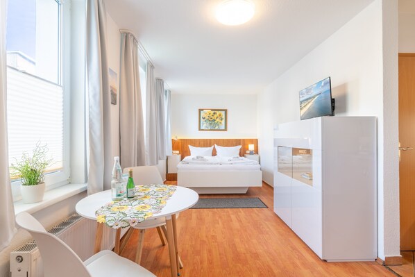 Ein kleines Hotelzimmer mit Bett, einem Tisch mit floralem Tischläufer, einem Kühlschrank und einem Fernseher auf einem Standfuß.