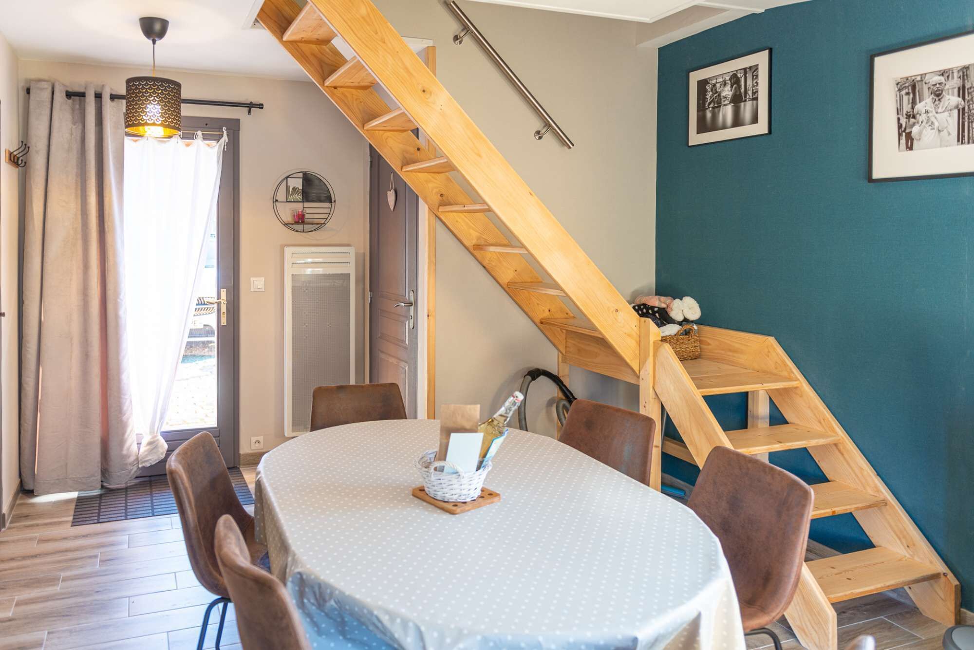 A dining area with a round table, chairs, a wooden staircase, and a blue wall with framed pictures.