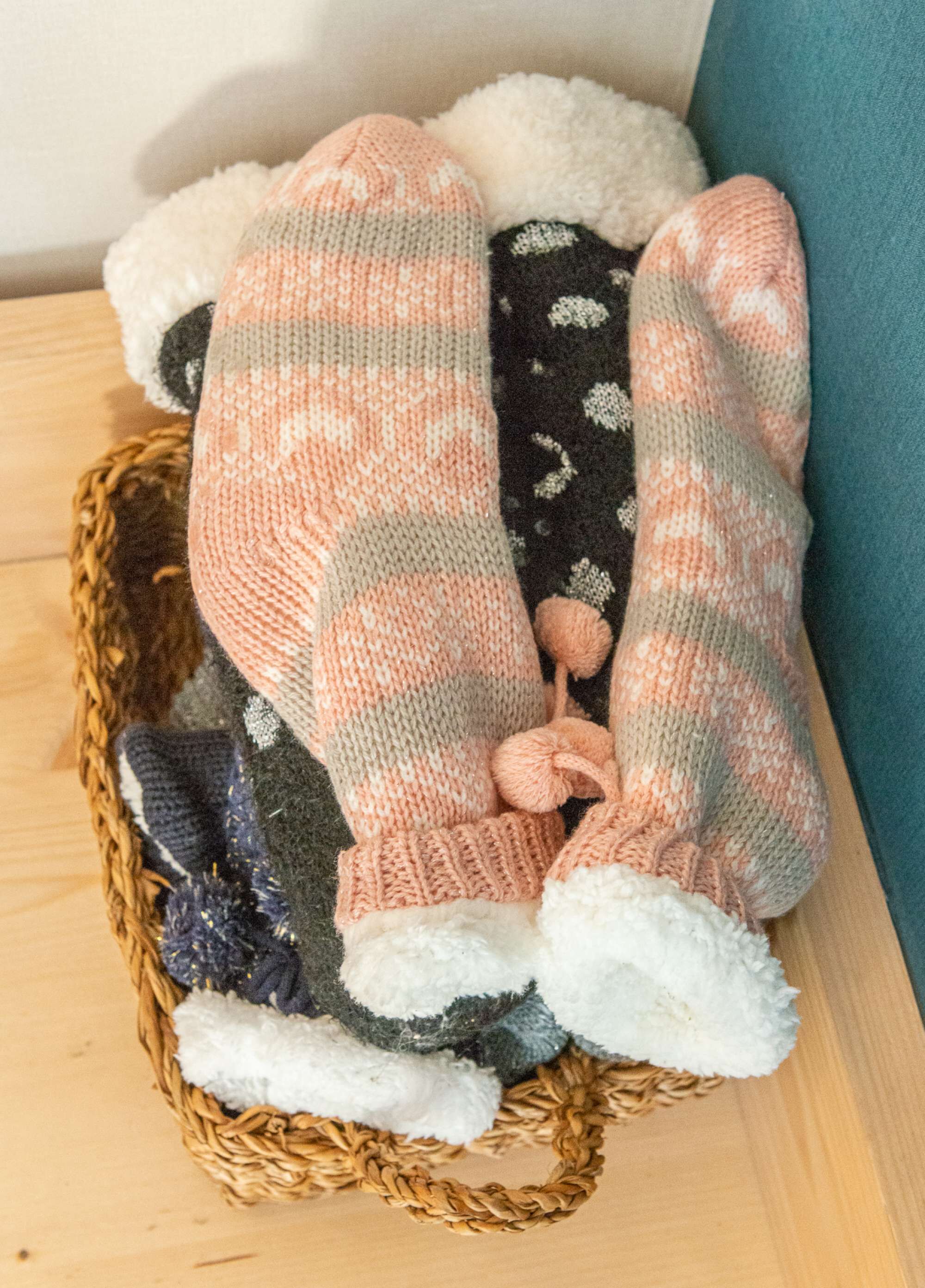 Ein Korb mit einem Paar gestrickter Socken mit Karomuster und Pompons, platziert auf einer Holzfläche neben einem blauen Kissen.