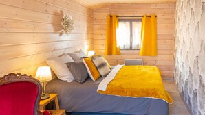 A wooden cabin bedroom with a bed, pillows, a chair, a table, and a window with curtains.