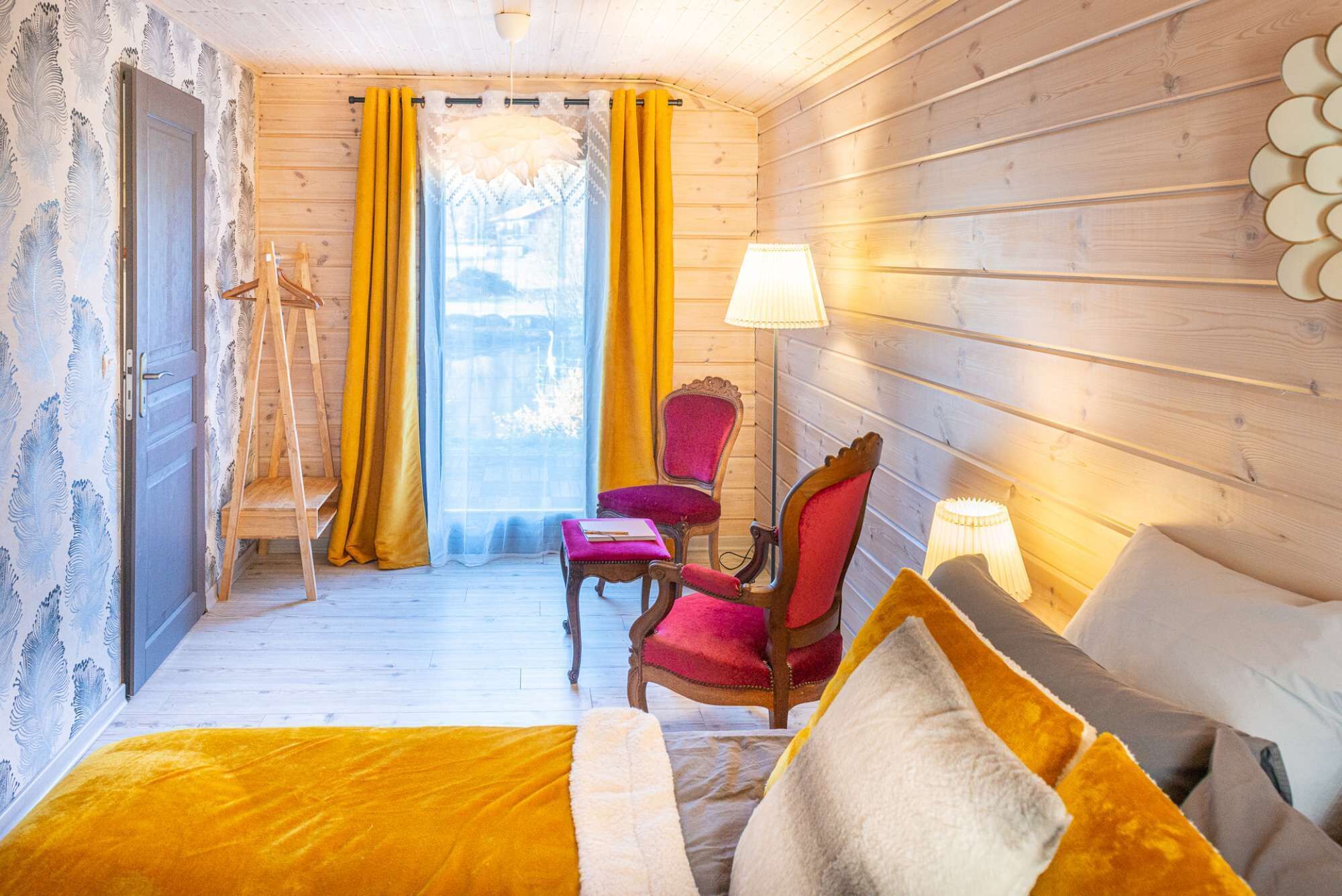 Une pièce chaleureuse aux murs en bois, avec un lit recouvert d’une couverture jaune, deux chaises et une lampe.