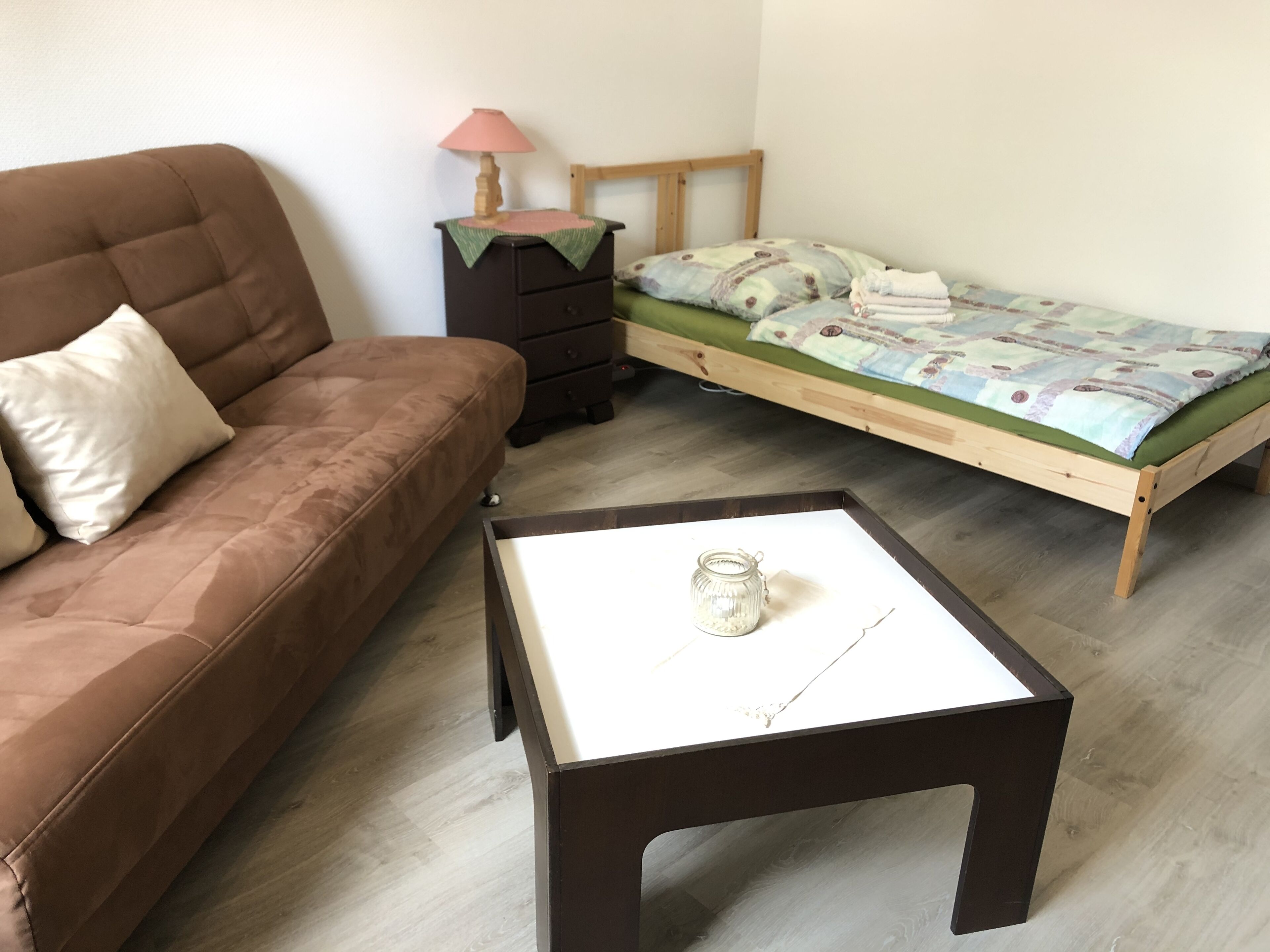 Quarto com um sofá marrom, uma cama de madeira com colchão verde, uma mesinha de cabeceira de madeira com abajur e uma mesa branca com um pote de vidro.