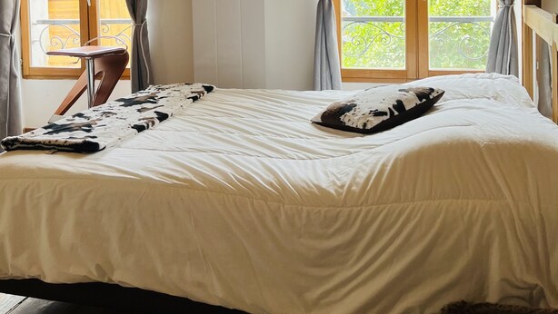 Ein Schlafzimmer mit einem großen Bett, einem Holzkopfteil, einem Nachttisch und Blick auf Grün durch ein Fenster.