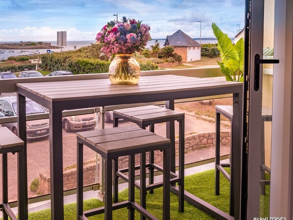 Un balcón con una mesa y sillas, un jarrón de flores y vistas a la costa con coches aparcados y un edificio al fondo.
