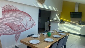 A dining area with a large fish mural, a wooden table set for six, and a kitchen in the background.