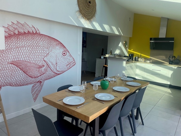 A dining area with a large fish mural, a wooden table set for six, and a kitchen in the background.