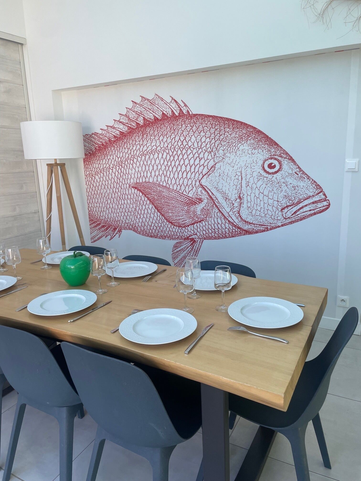 Una mesa de comedor dispuesta con platos, cubiertos y vasos, con un gran mural de un pez en la pared de fondo.