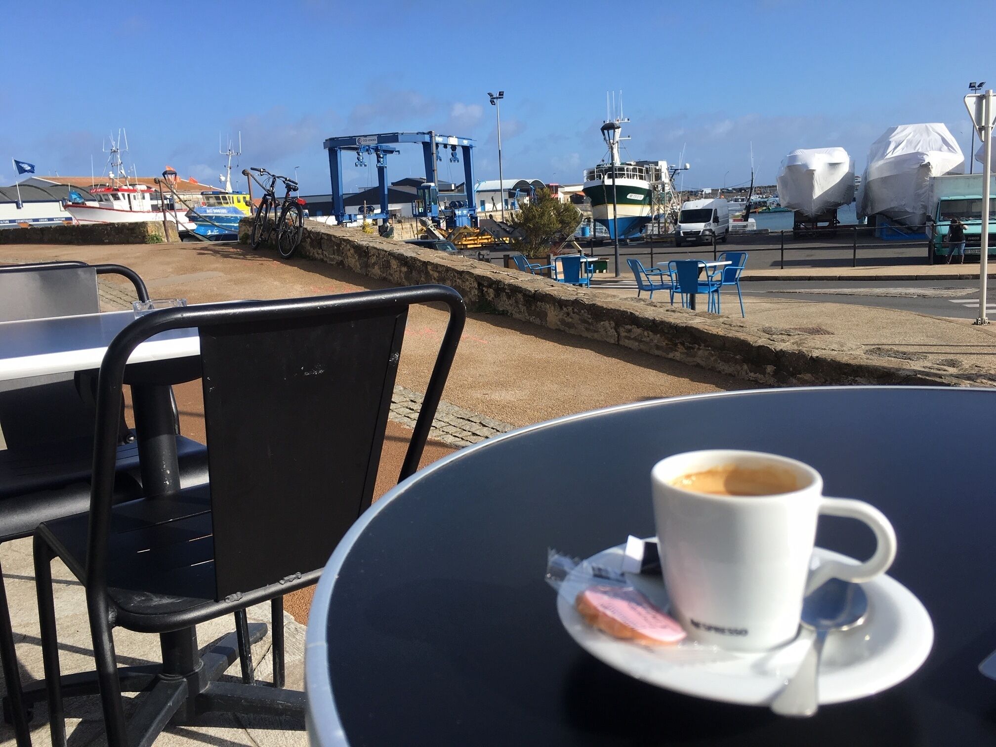 A table with a cup of coffee and a pastry, overlooking a harbor with boats and a crane.