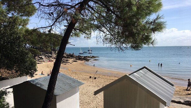 A beach with two small white wooden structures, likely beach huts, and a clear view of the sea with boats in the distance.