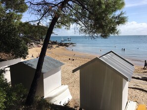 Una playa con dos pequeñas construcciones de madera blanca, probablemente cabañas de playa, y una vista despejada del mar con barcos a lo lejos.