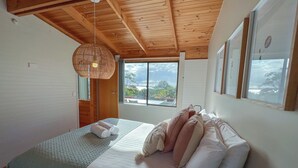 A bedroom with a wooden ceiling, a large bed with pillows, a hanging light fixture, and a window with a view. - HEXAGON BEACH HOUSE - Great views of the Bay of Fires (Binalong Bay)