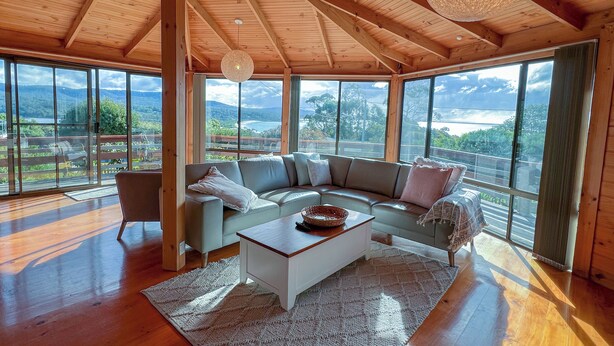 A spacious living room with a large sectional sofa, a coffee table, and a view of a mountainous landscape through large windows. - HEXAGON BEACH HOUSE - Great views of the Bay of Fires (Binalong Bay)