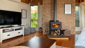 A living room with a TV, white entertainment unit, and a brick fireplace with logs. - HEXAGON BEACH HOUSE - Great views of the Bay of Fires (Binalong Bay)