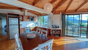 A spacious kitchen with wooden cabinets, a dining area with a wooden table and white chairs, and a large sliding glass door leading to a scenic outdoor view. - HEXAGON BEACH HOUSE - Great views of the Bay of Fires (Binalong Bay)