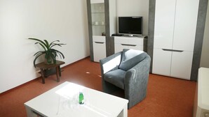 A modern living room with a grey sofa, a white coffee table, a small table with a plant, a TV on a cabinet, and a wardrobe.