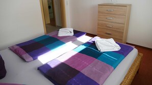 A neatly made bed with a colorful checkered bedspread and two folded white towels. A wooden dresser with drawers is to the right of the bed.