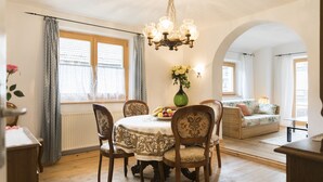 Un coin repas avec une table ronde, des chaises et un lustre. On aperçoit un canapé et une fenêtre avec des rideaux à l’arrière-plan.