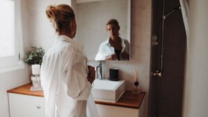 Une personne vêtue d’une robe blanche se tient dans une salle de bain, devant un miroir, un comptoir en bois et un meuble blanc.