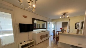 A modern living room with a flat-screen TV, a white console table, a sofa, and a dining area with chairs. - Spacious apartment for 4, balcony, sea view, near beach, parking, cleaning included (Saint-Hilaire-de-Riez)