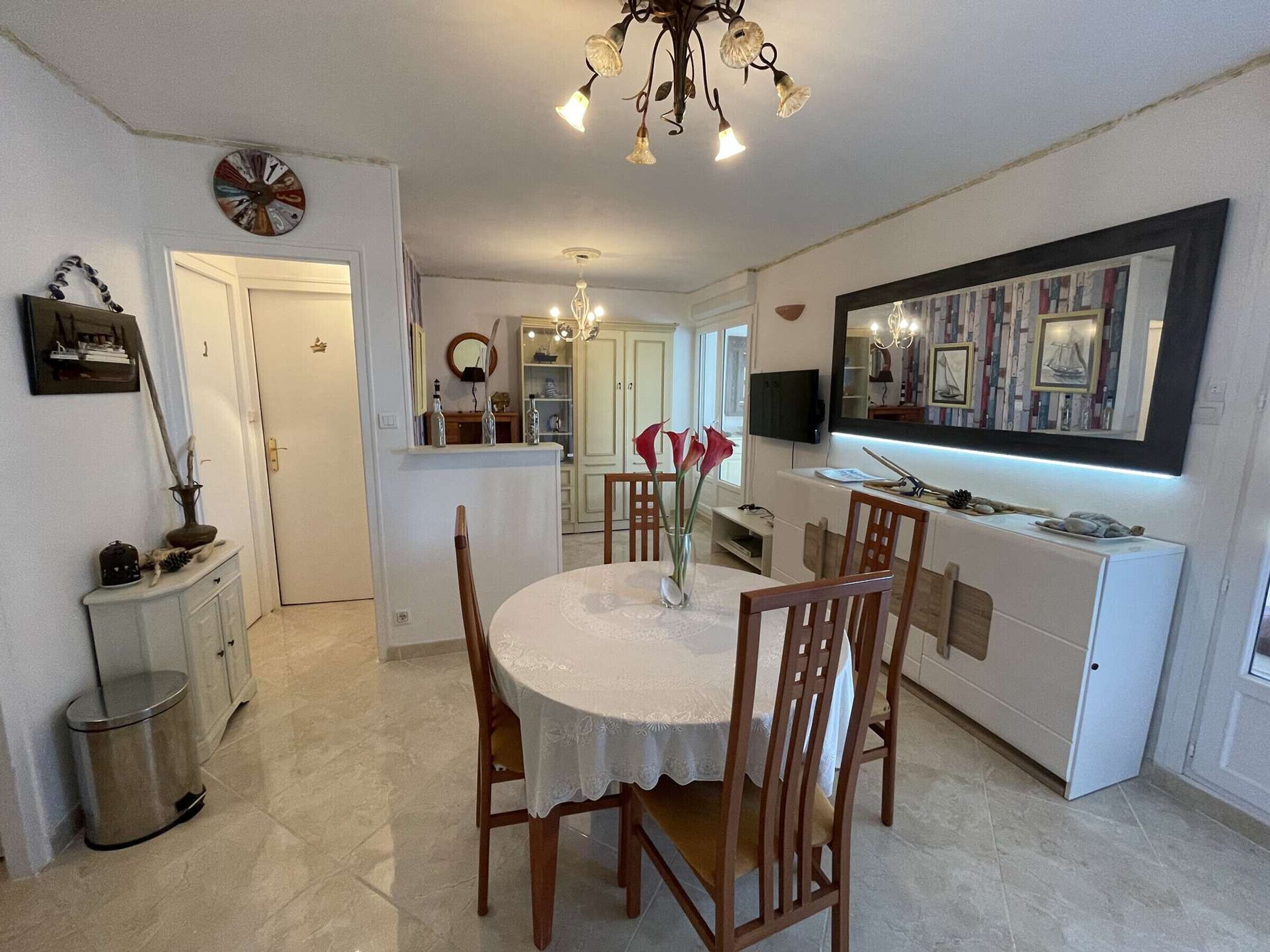 A dining area with a round table, chairs, a chandelier, a clock, and a mirror on the wall.