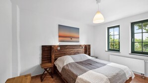 Een slaapkamer met een houten hoofdbord, een bed met een quilt, een nachtkastje, een wandlamp, een raam met uitzicht op groen, en een schilderij van een zonsondergang.