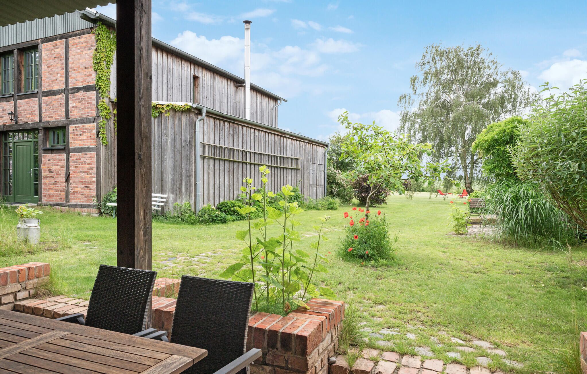 Un bâtiment rustique avec une façade en briques et des poutres en bois, une cour verdoyante avec un chemin en briques et un espace extérieur aménagé avec des chaises en osier.