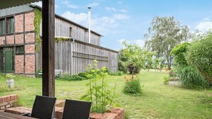 Een landelijk gebouw met een bakstenen gevel en houten balken, een gazon met een metselpad, en buitenzitplaatsen met rieten stoelen.