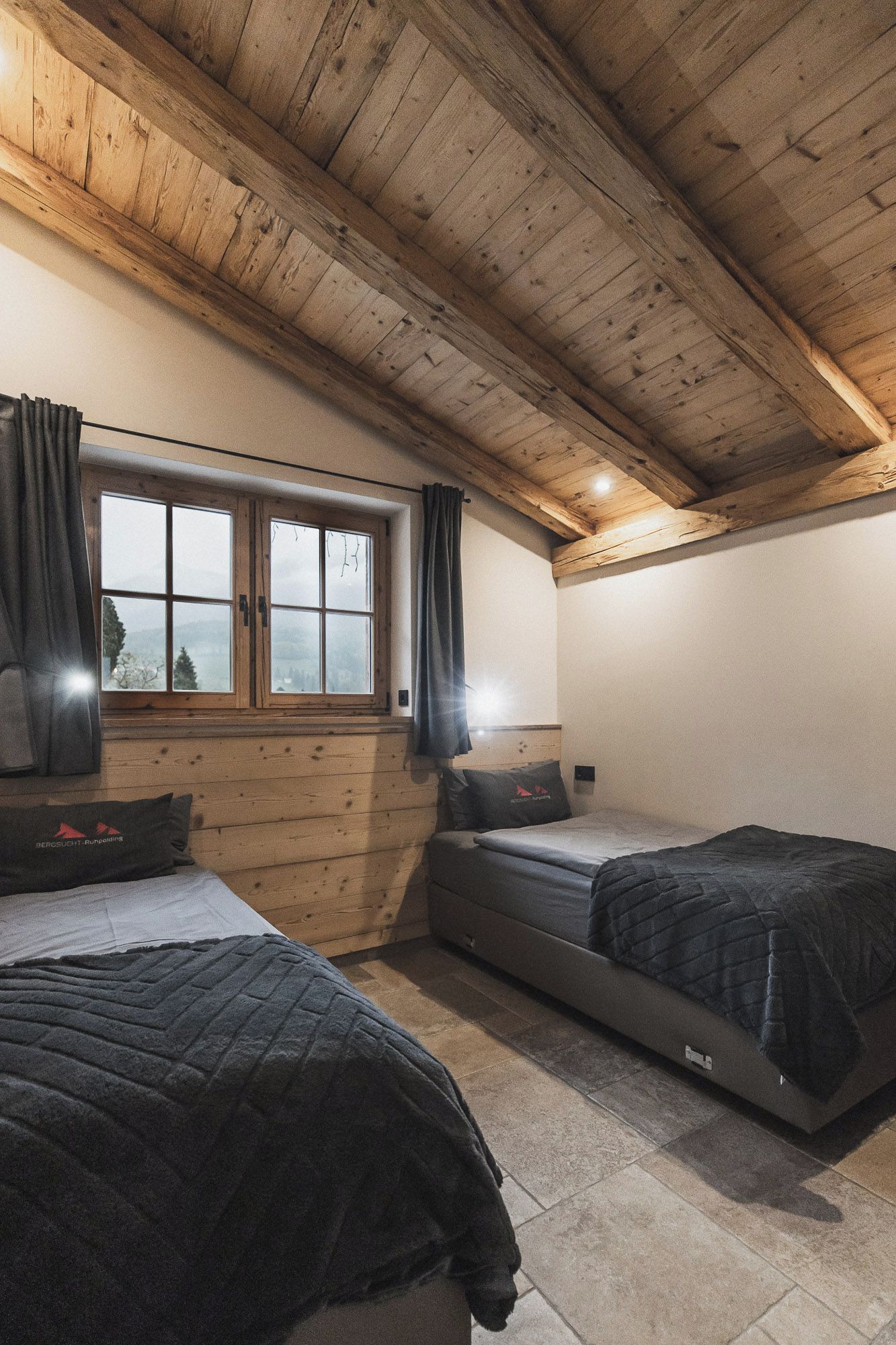 Une pièce avec des poutres apparentes en bois au plafond, une fenêtre avec des rideaux, deux lits avec une literie sombre et une vue sur l’extérieur.