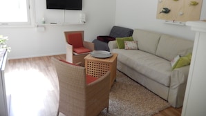 A living room with a sofa, a chair, a flat-screen TV, and a wall-mounted artwork. - Ferienhaus Waldoase für 2 - 3 Personen, Freistehend auf Eingezäuntem Grundstück (Beelitz)